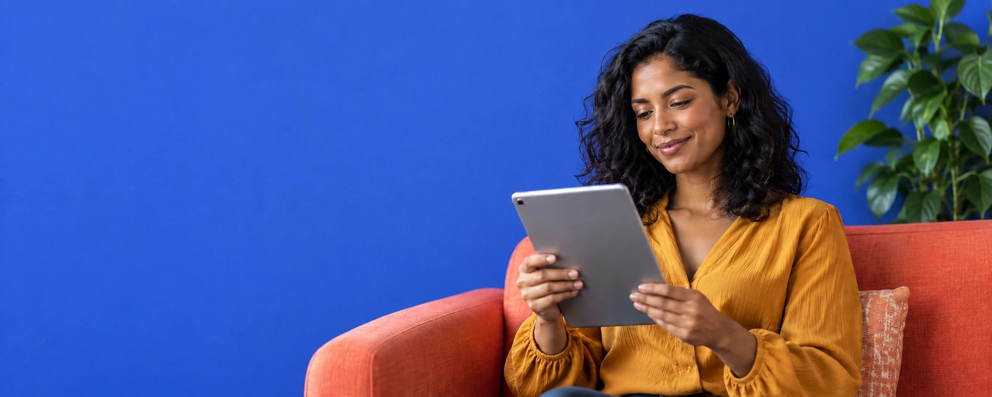 Mulher de blusa âmbar sentada em sofá coral, usando um tablet diante de parede azul e planta verde à direita.